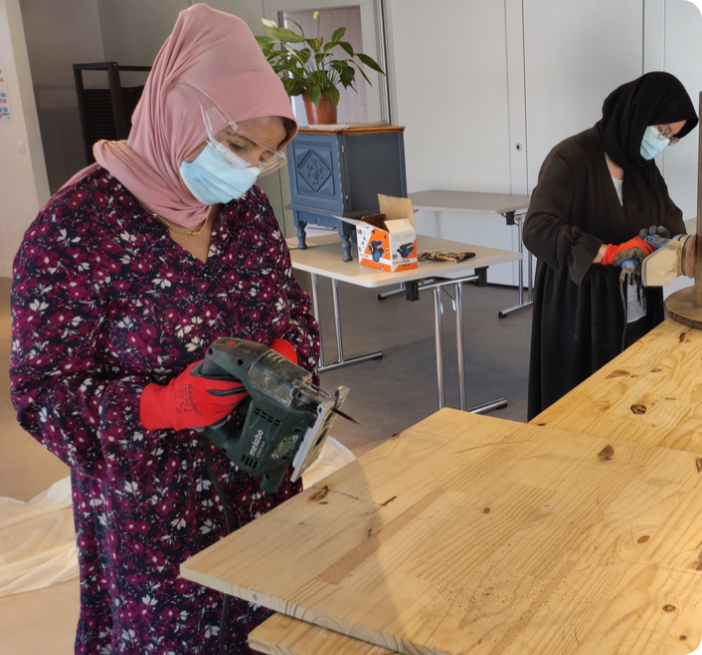 Atelier de l’association : deux participantes découpent et poncent des planches de bois, gants et lunettes de protection sur le nez.