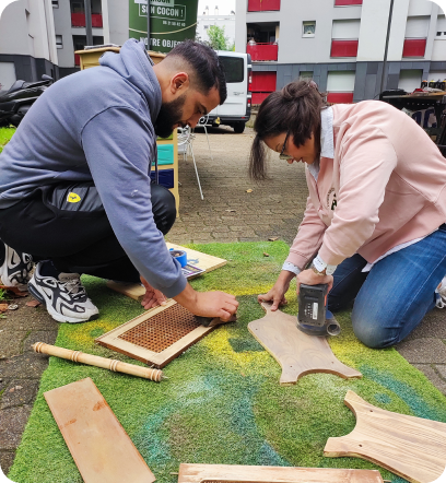 Deux bénévoles poncent des pieds et des planches de meuble en bois, en extérieur, au pied d’un immeuble
