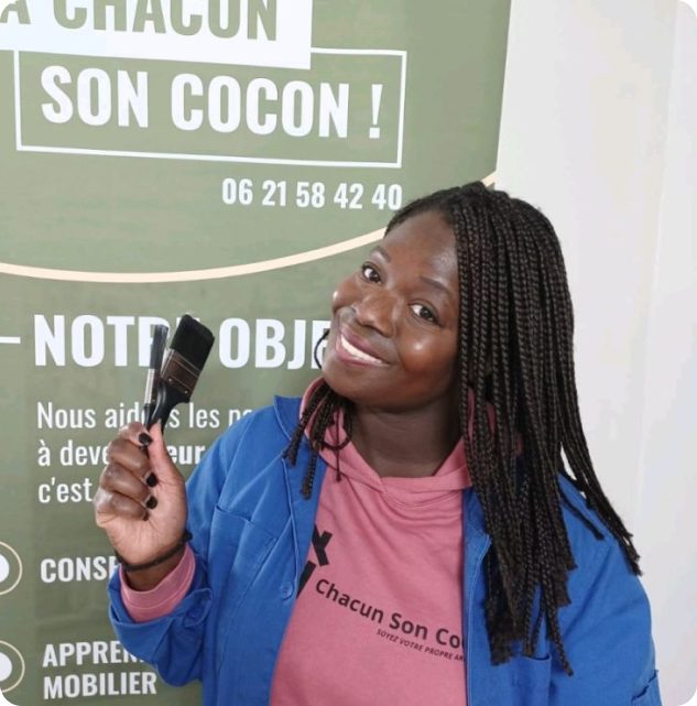 Assetou Coulibaly, souriante, deux pinceaux à la main devant le kakémono de l’association