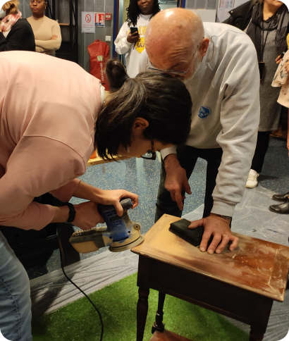 Deux bénévoles poncent une petite table en bois avec une ponceuse électrique pendant un atelier