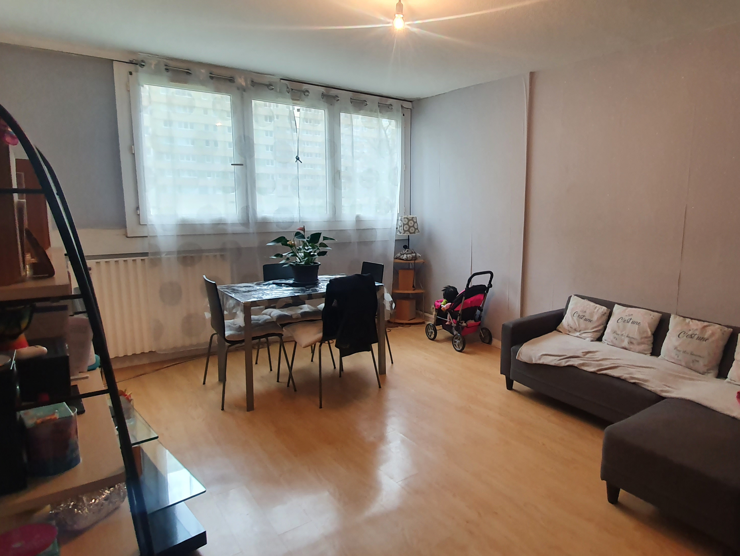 Avant : le séjour avec la table à manger et ses chaises au centre de la pièce, le canapé contre le mur du fond et une poussette près de la fenêtre